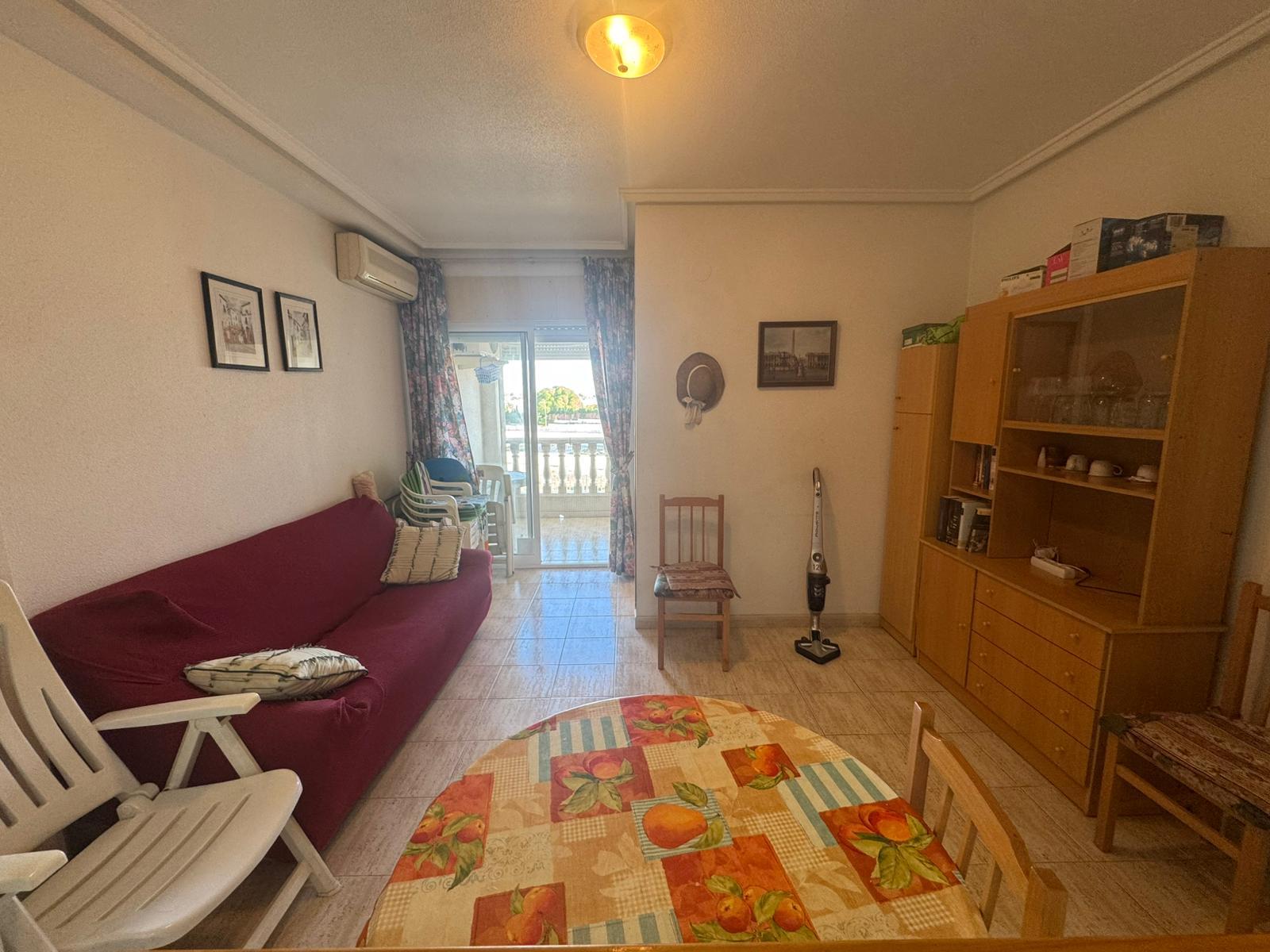 Living room with a maroon sofa, white folding chairs, and a wooden cabinet; floral curtains frame a balcony door beyond.