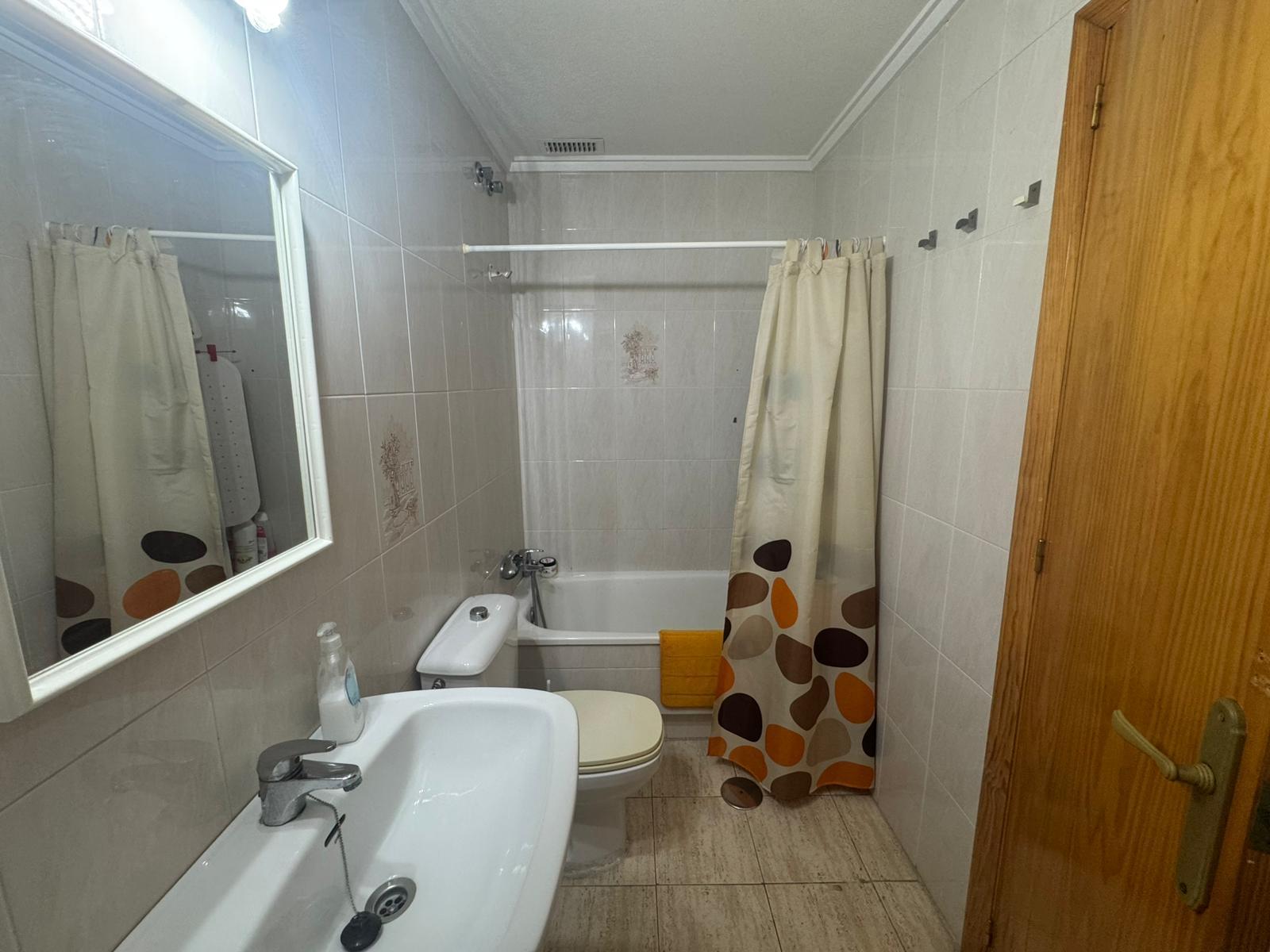 Small bathroom with white sink and toilet, bathtub with beige shower curtain featuring orange/brown circles, and a wooden door left.