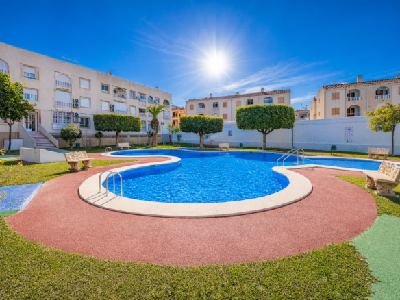 Sunlit courtyard pool area with a curving blue swimming pool, red walking paths, green lawns, and beige apartment buildings surrounding it.