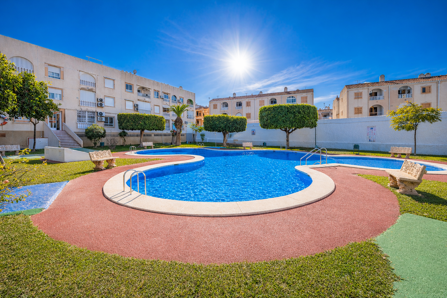 Sunlit courtyard pool area with a curving blue swimming pool, red walking paths, green lawns, and beige apartment buildings surrounding it.