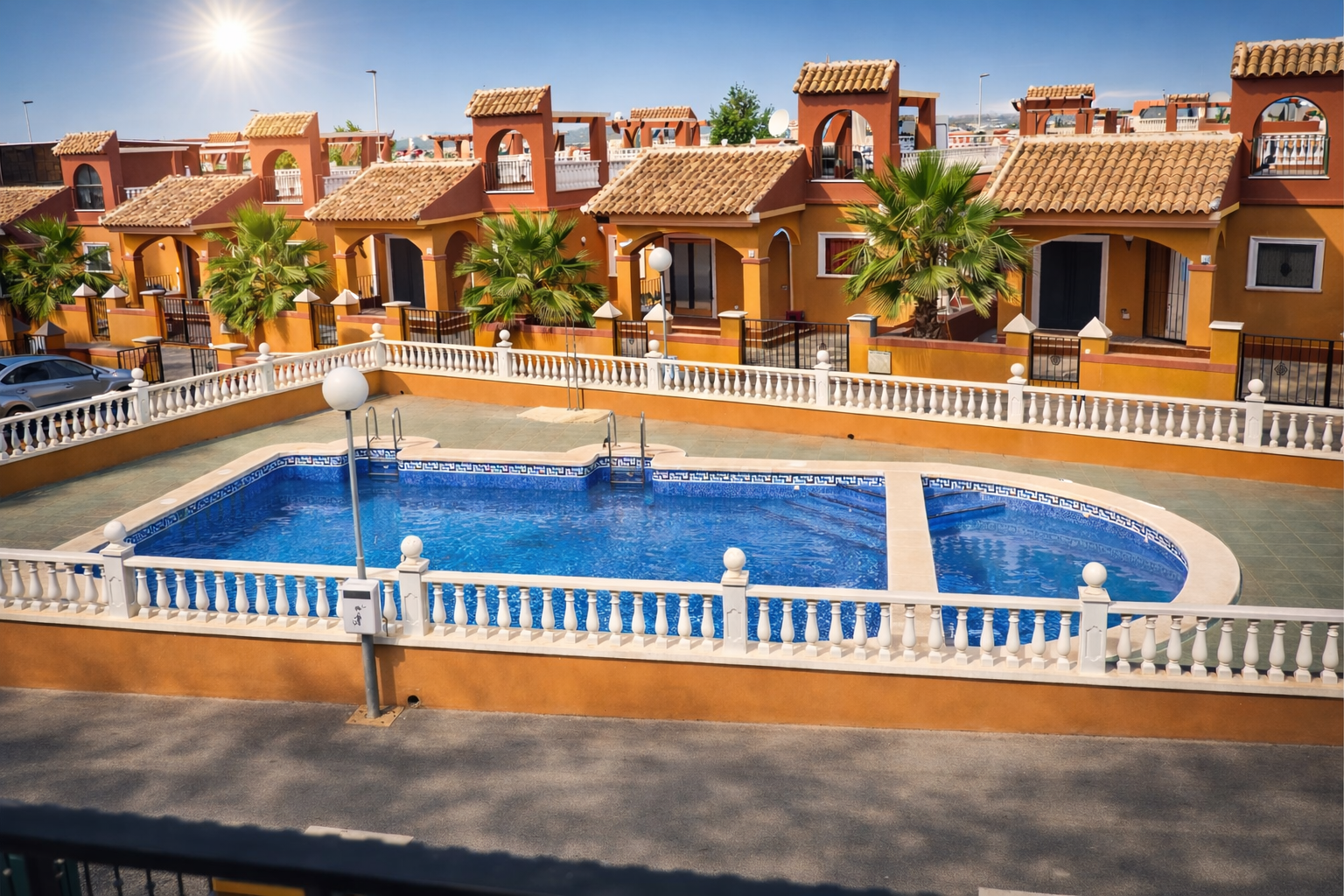 Une piscine commune extérieure entourée de maisons de style méditerranéen orange et de balustrades blanches, sous un ciel radieux.