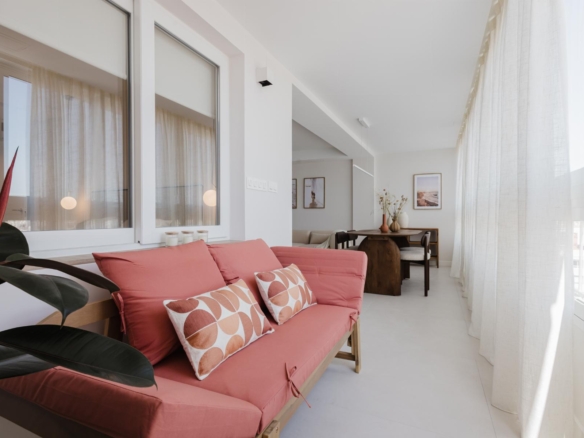 Bright living room with a coral sofa and geometric-pattern pillows, large windows with sheer white curtains, and a dining area in the background.