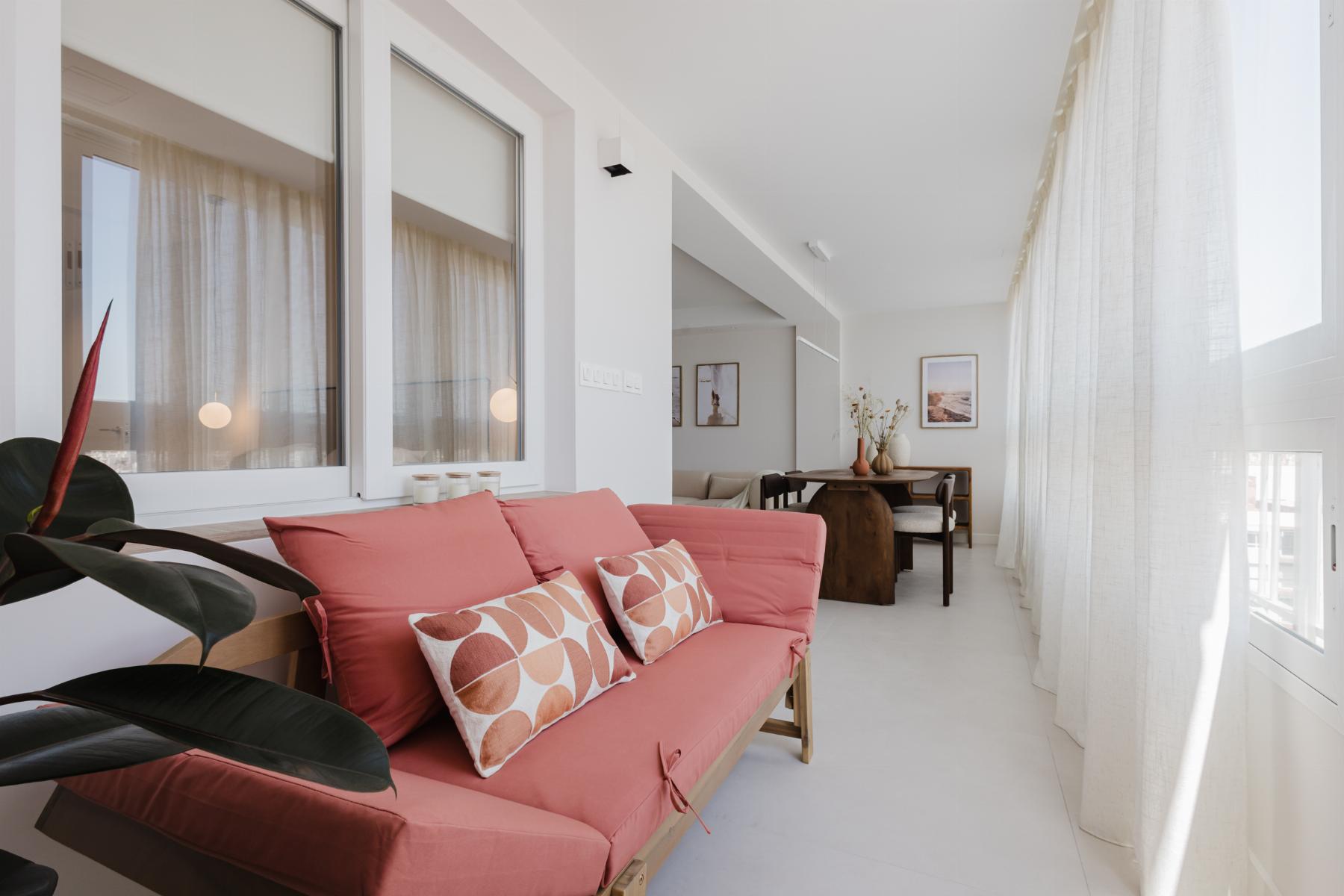 Bright living room with a coral sofa and geometric-pattern pillows, large windows with sheer white curtains, and a dining area in the background.