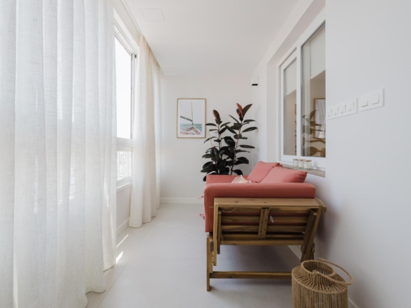 Bright balcony seating area with a pink cushion sofa, white curtains, and a tall plant beside a framed wall art piece