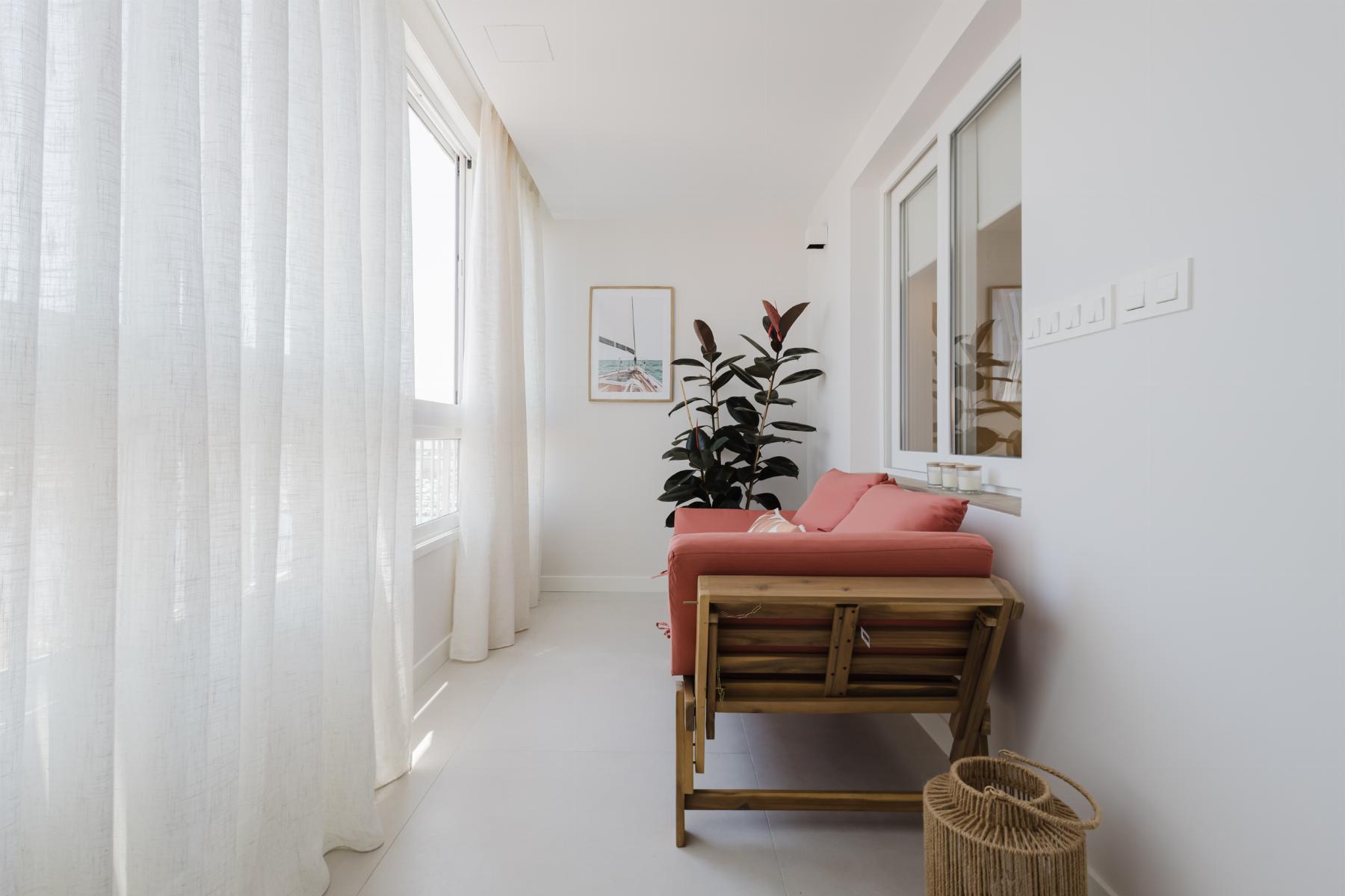Bright balcony seating area with a pink cushion sofa, white curtains, and a tall plant beside a framed wall art piece