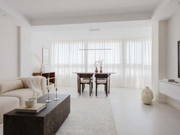 Bright minimalist living and dining area with a beige sofa, dark marble coffee table, and a wooden dining table by large windows with sheer curtains.