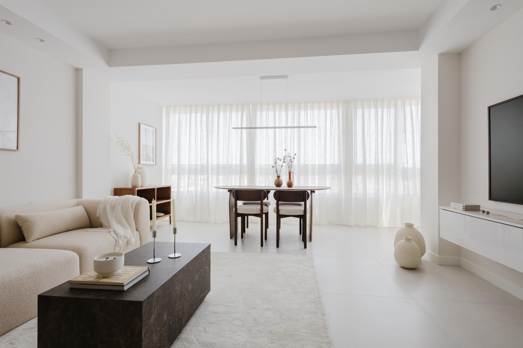 Bright minimalist living and dining area with a beige sofa, dark marble coffee table, and a wooden dining table by large windows with sheer curtains.