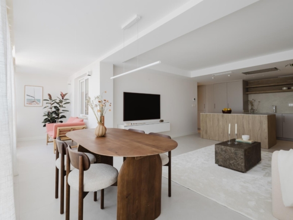 Bright open-plan living area with wooden dining table, gray upholstered chairs, potted plant, and a wall-mounted TV.