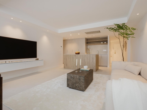Open-concept living room with a wall-mounted flat-screen TV above a white console, beige sofa to the right, and a light, neutral color palette; kitchen area visible in the background.