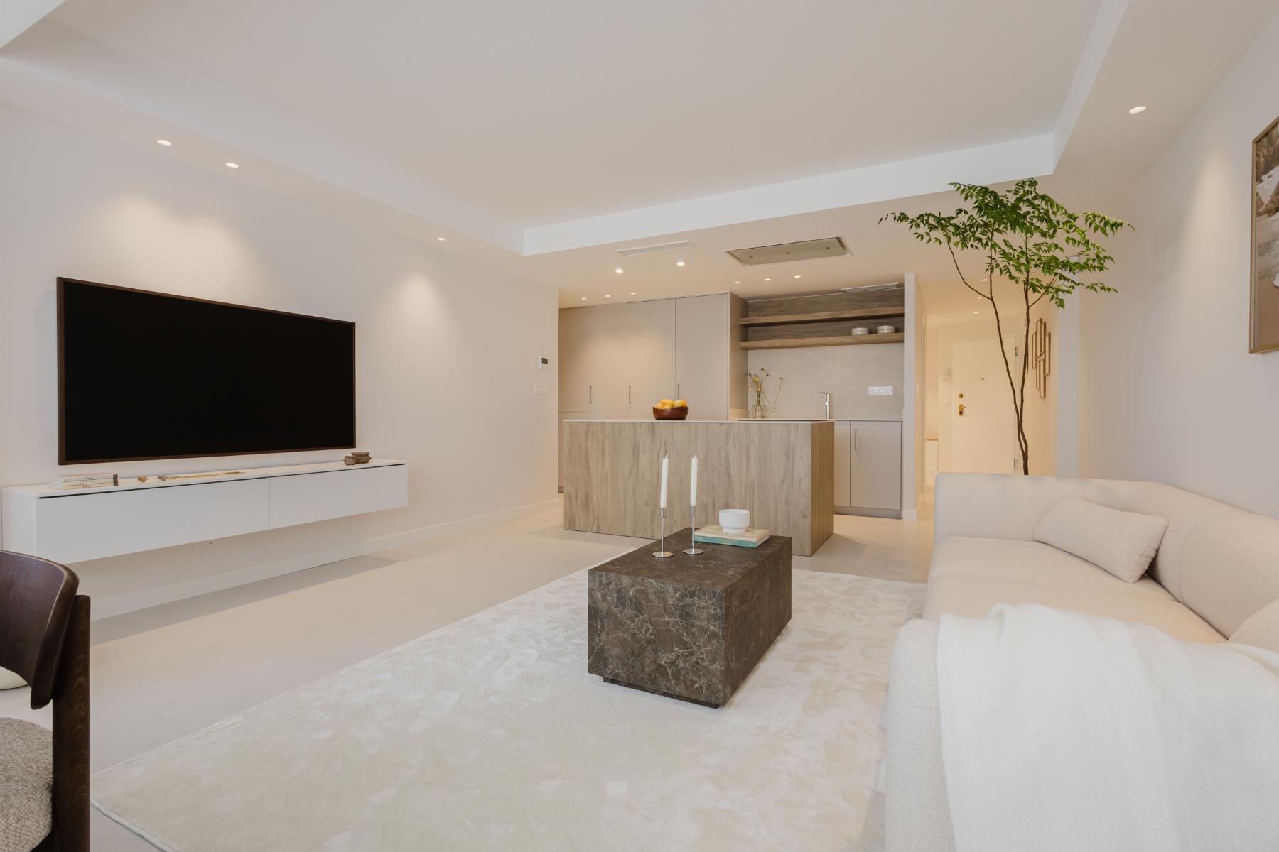 Open-concept living room with a wall-mounted flat-screen TV above a white console, beige sofa to the right, and a light, neutral color palette; kitchen area visible in the background.
