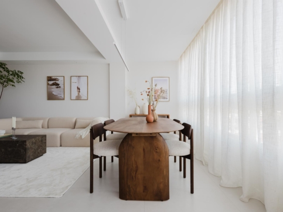 Bright open living and dining area with a long wooden table, beige chairs, a cream sectional sofa, and sheer white curtains along the right wall.