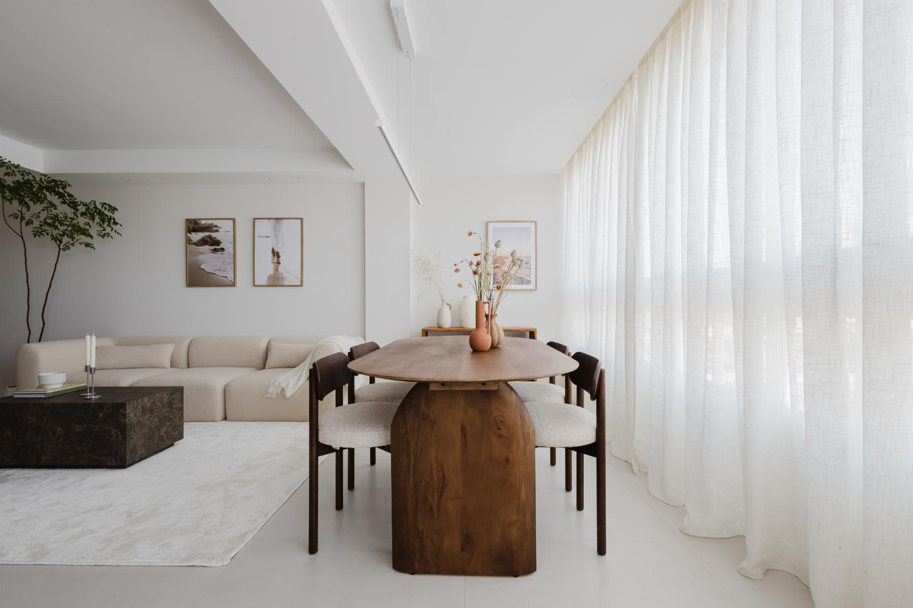 Bright open living and dining area with a long wooden table, beige chairs, a cream sectional sofa, and sheer white curtains along the right wall.