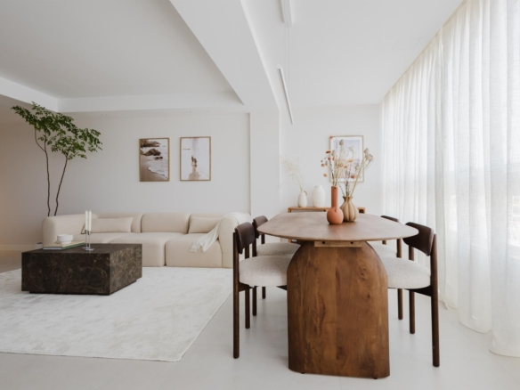 Bright open-plan living and dining room with a beige sectional sofa, a dark marble coffee table, and a round wooden dining table with upholstered chairs under soft natural light from sheer white curtains