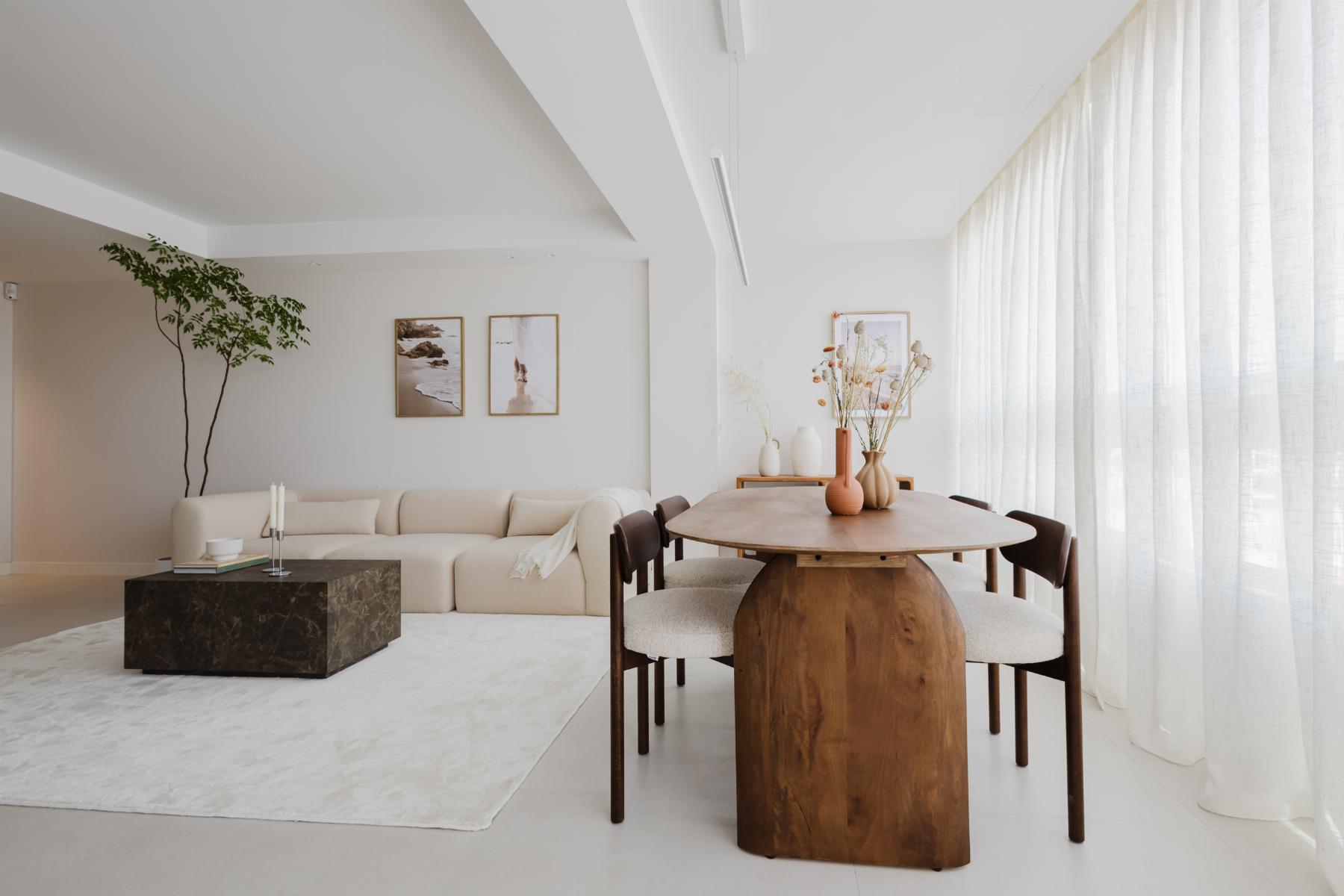 Bright open-plan living and dining room with a beige sectional sofa, a dark marble coffee table, and a round wooden dining table with upholstered chairs under soft natural light from sheer white curtains