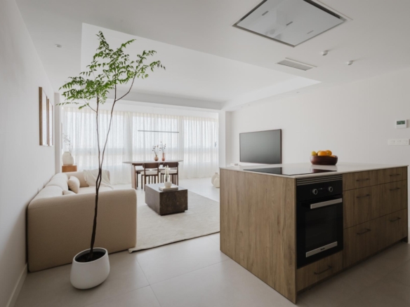 Bright open-concept living area and kitchen with a wooden island, beige sofa, and a tall potted tree near large windows with sheer white curtains.