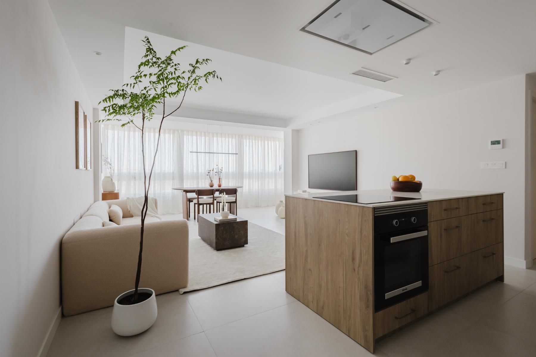 Bright open-concept living area and kitchen with a wooden island, beige sofa, and a tall potted tree near large windows with sheer white curtains.