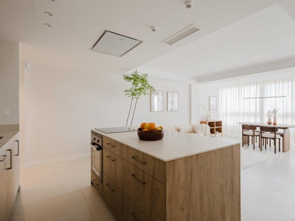 Bright open-concept kitchen with a wooden island, fruit bowl, and adjacent dining area by large windows with sheer curtains.