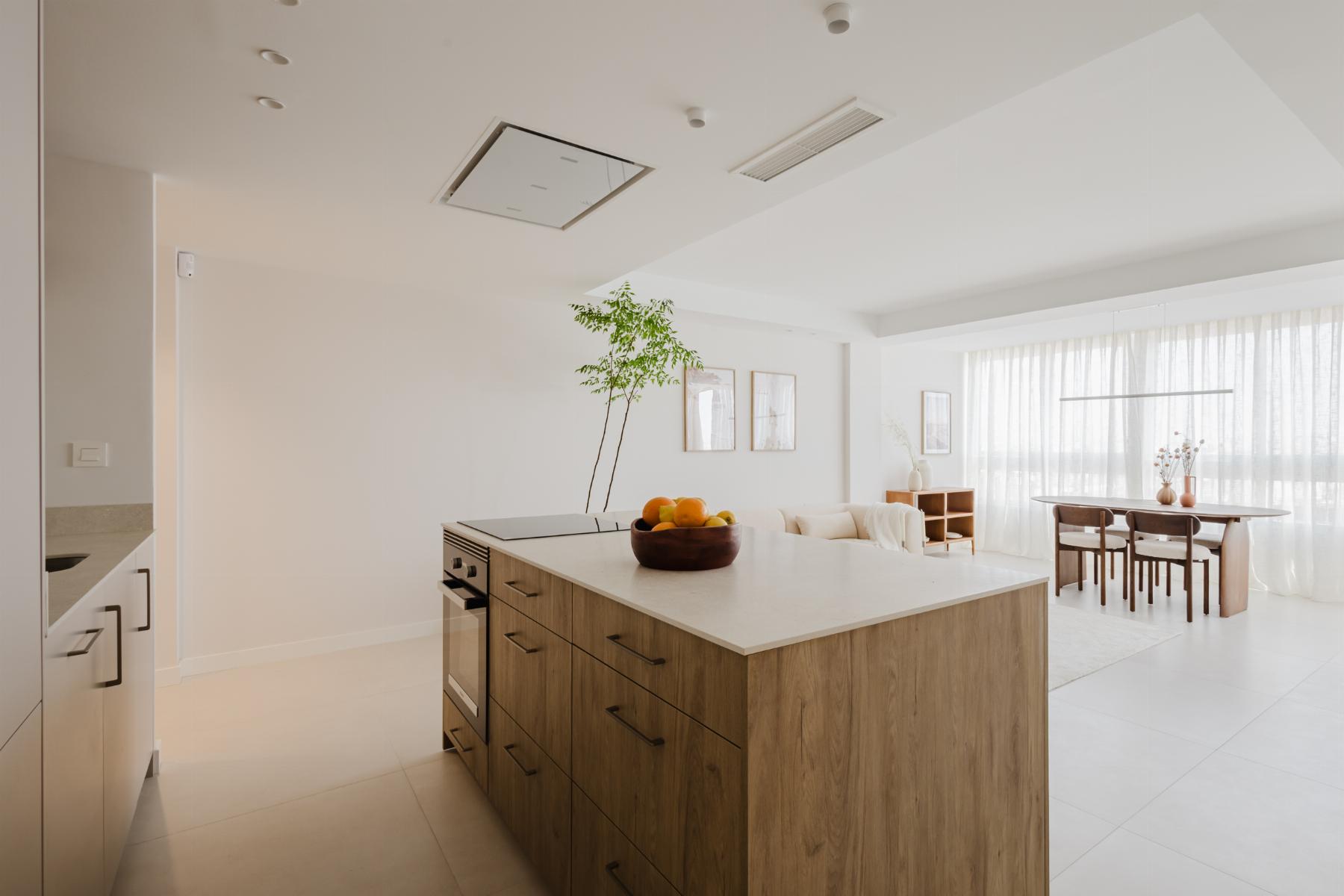 Bright open-concept kitchen with a wooden island, fruit bowl, and adjacent dining area by large windows with sheer curtains.