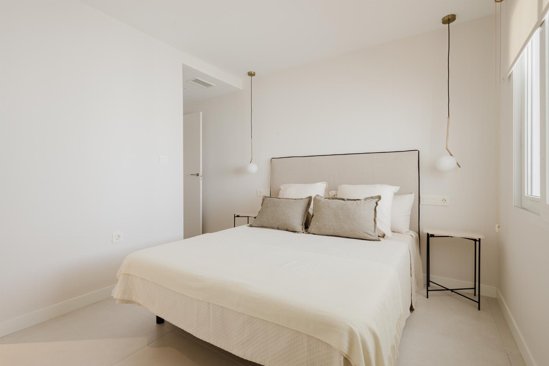 Minimalist white bedroom with a beige upholstered headboard bed, white bedding, and two hanging pendant lights by each side.
