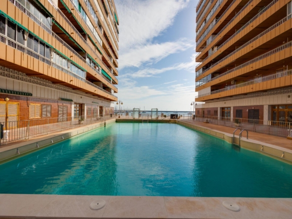 Rectangular pool between two tall orange apartment buildings with balconies, view of the sea in the distance.