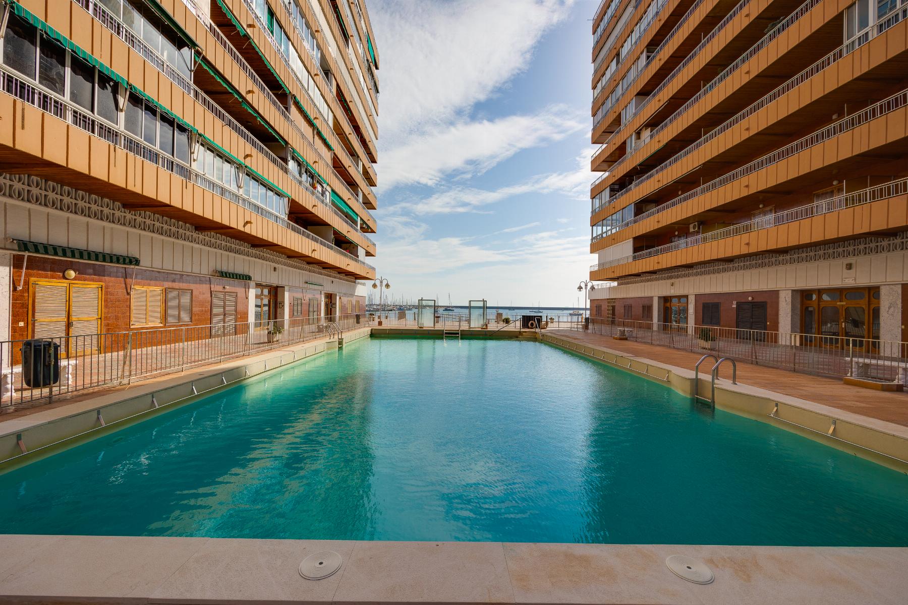 Rectangular pool between two tall orange apartment buildings with balconies, view of the sea in the distance.