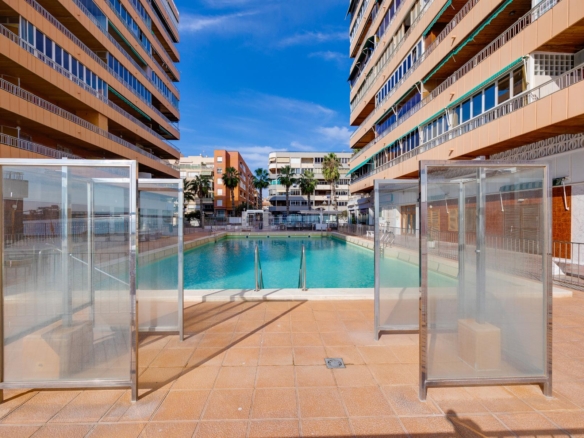 Outdoor swimming pool framed by tall beige apartment buildings with balconies and glass windbreaks, blue sky above and palm trees in the background.