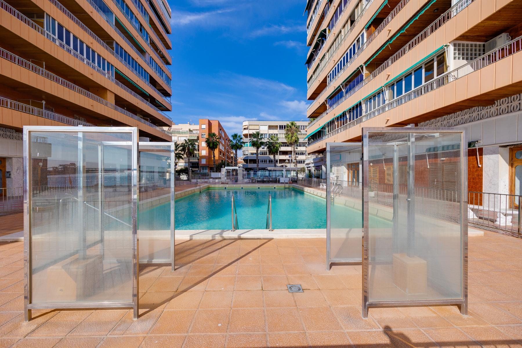 Outdoor swimming pool framed by tall beige apartment buildings with balconies and glass windbreaks, blue sky above and palm trees in the background.