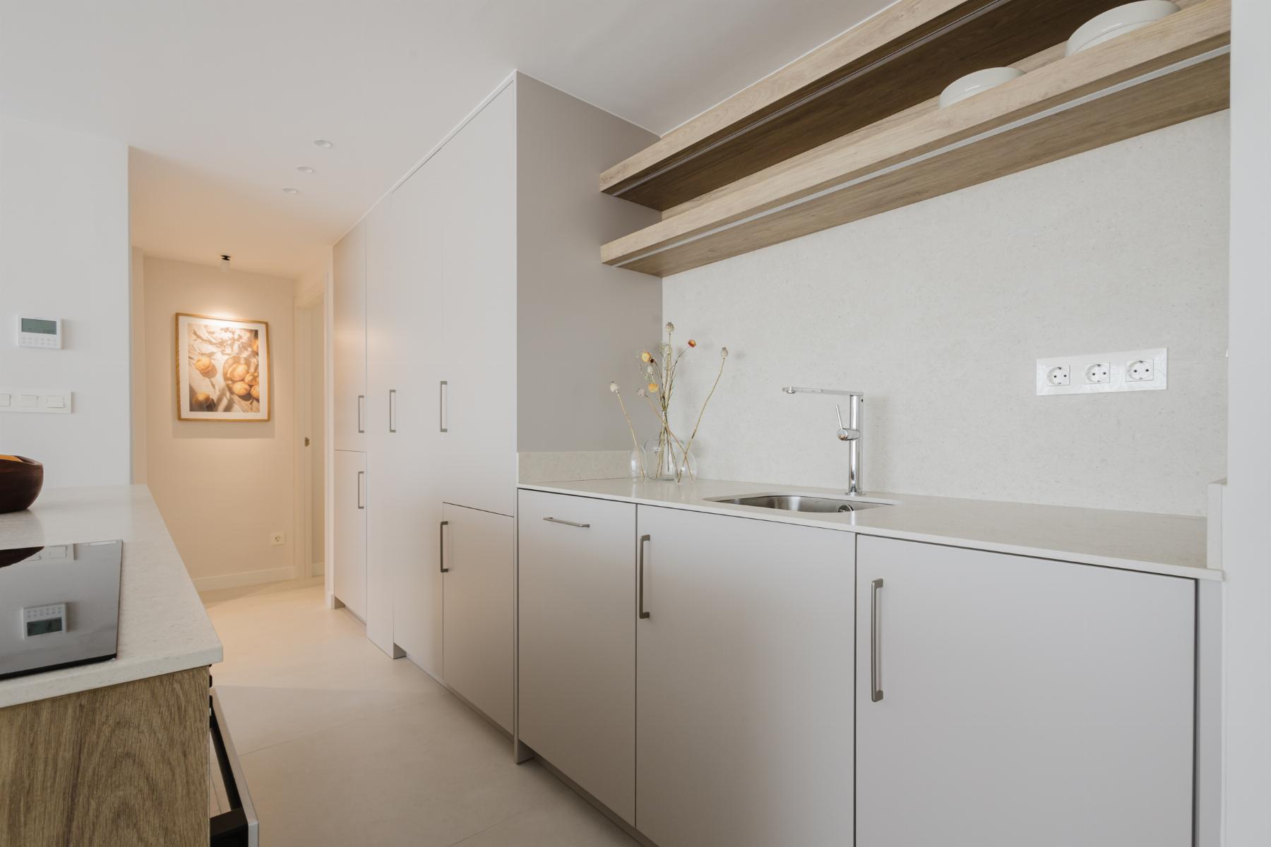 Modern white kitchen with light wood open shelves and a stainless sink area, hallway beyond with framed art