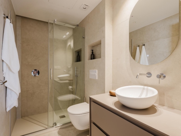 Modern beige-tiled bathroom with glass shower enclosure, round vessel sink on a grey vanity, and a large circular wall mirror.