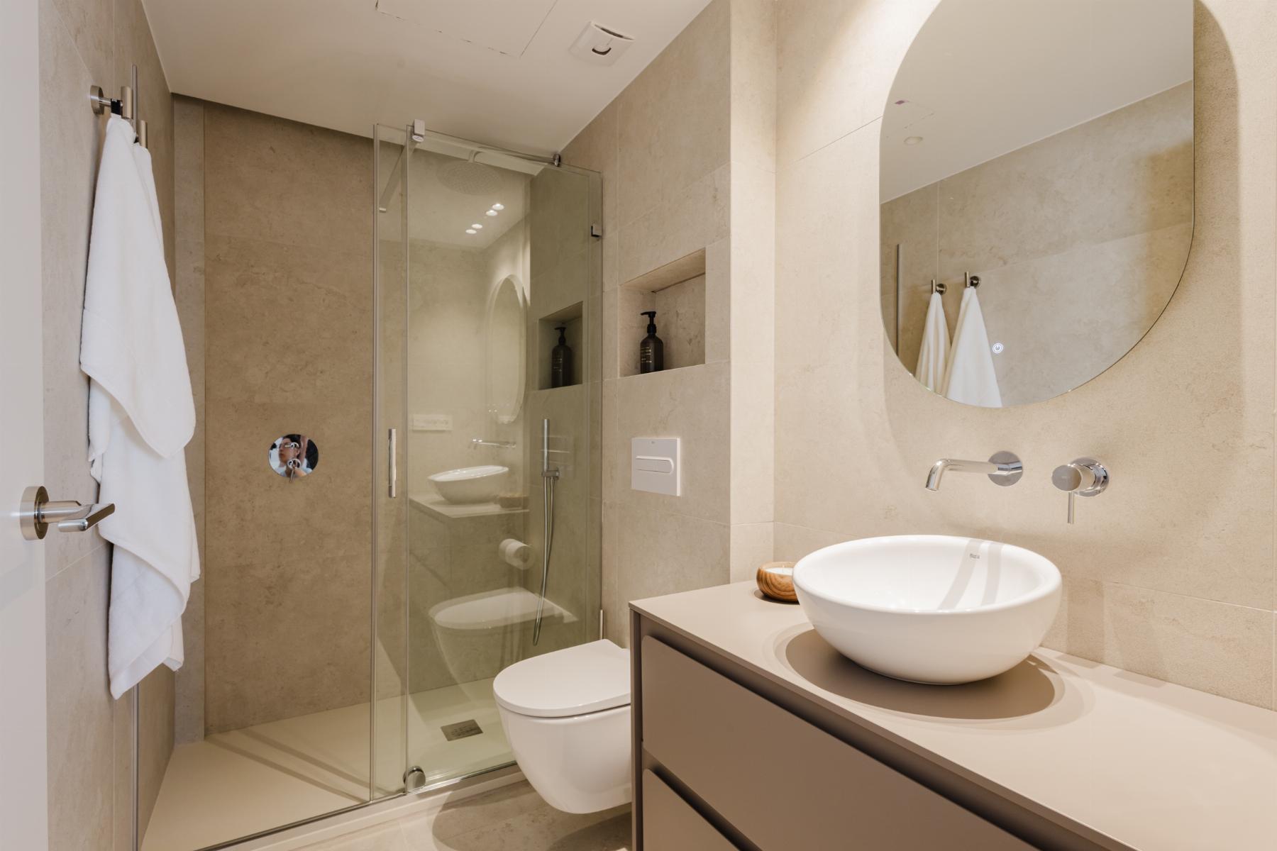 Modern beige-tiled bathroom with glass shower enclosure, round vessel sink on a grey vanity, and a large circular wall mirror.
