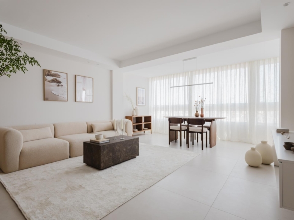 Bright minimalist living room with a beige sofa, marble coffee table and a dining area by large windows with sheer curtains.