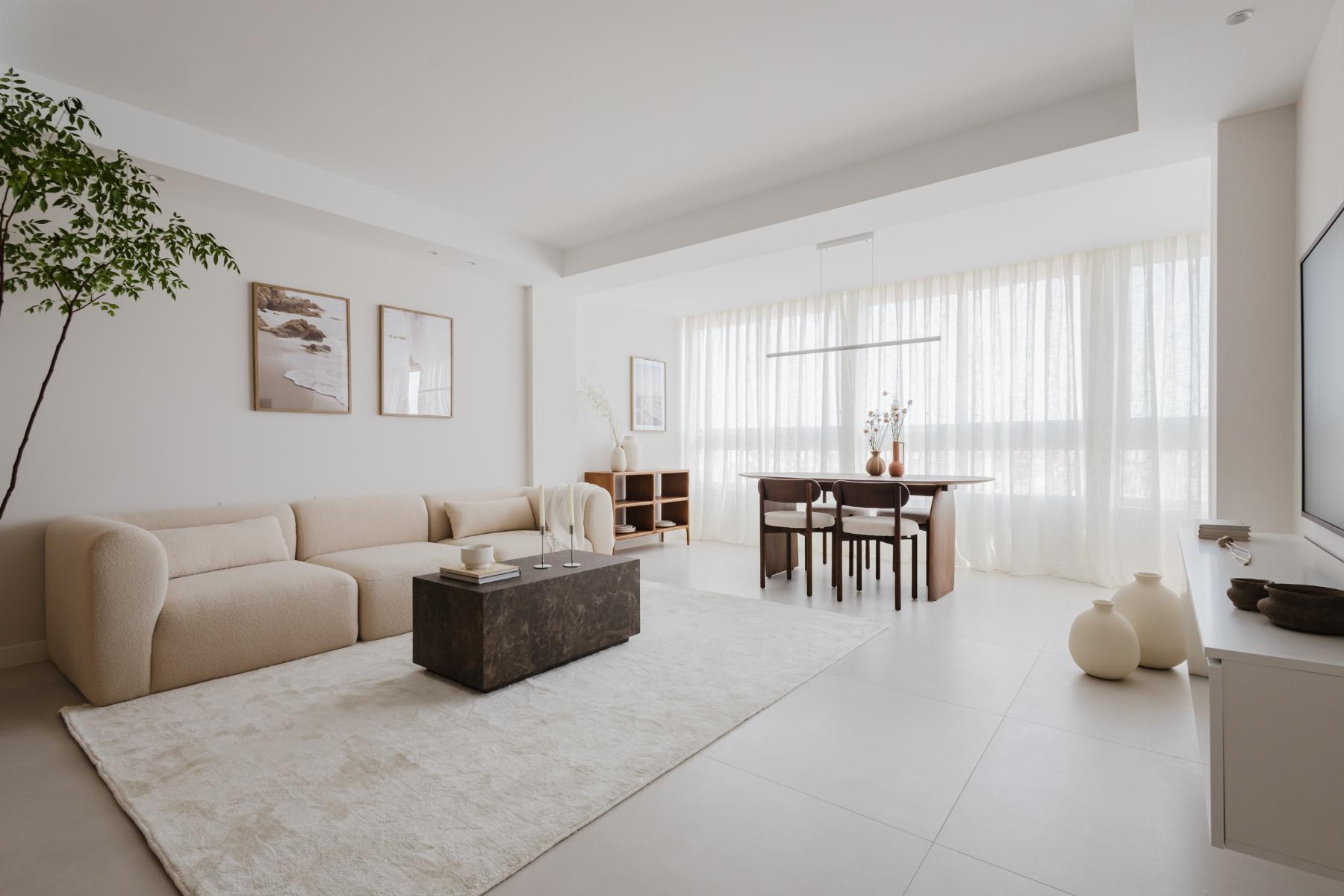 Bright minimalist living room with a beige sofa, marble coffee table and a dining area by large windows with sheer curtains.