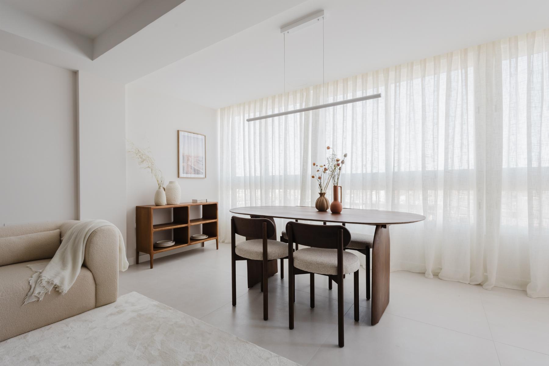 Bright minimalist dining area with a round wooden table, four dark chairs, and sheer white curtains