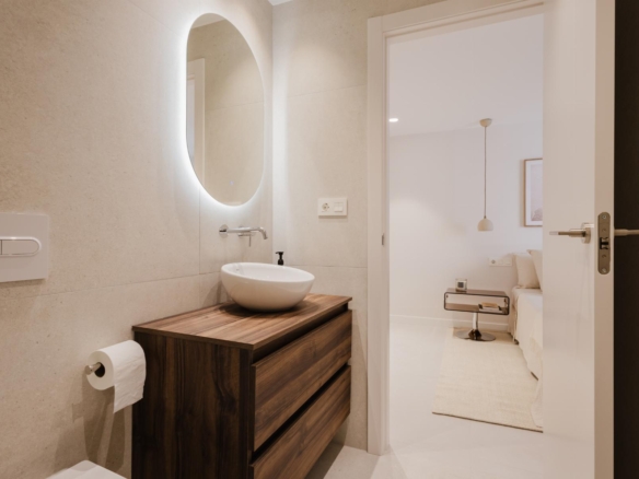 Modern bathroom with a circular white sink on a dark wooden vanity and a backlit oval mirror on beige tiled walls