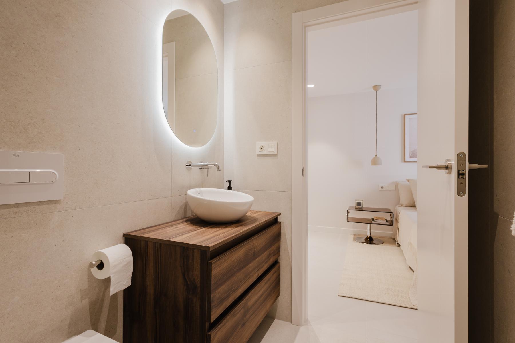 Modern bathroom with a circular white sink on a dark wooden vanity and a backlit oval mirror on beige tiled walls
