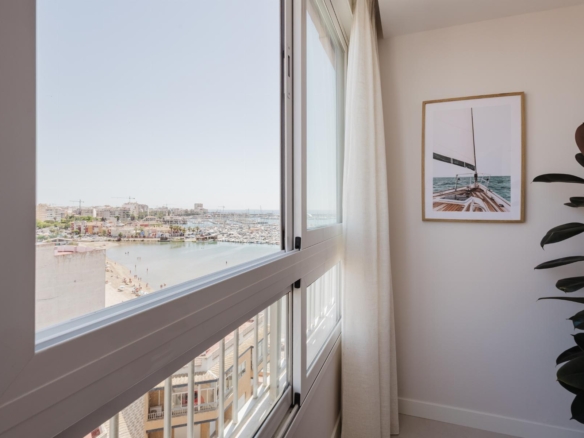 Interior corner with large window showing a marina and beach, white curtains, and a framed sailboat photo on the wall next to a potted plant.