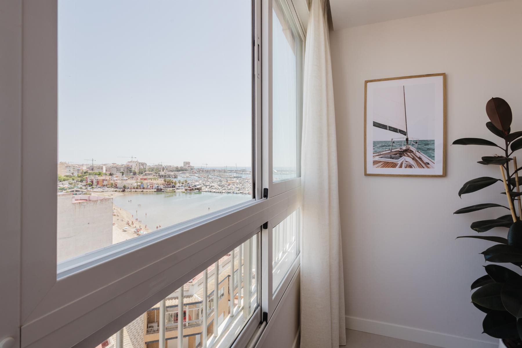 Interior corner with large window showing a marina and beach, white curtains, and a framed sailboat photo on the wall next to a potted plant.