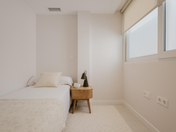 Minimal neutral bedroom with a single bed, a small wooden nightstand, black lamp, and a cup on the table beside a large window with blinds.