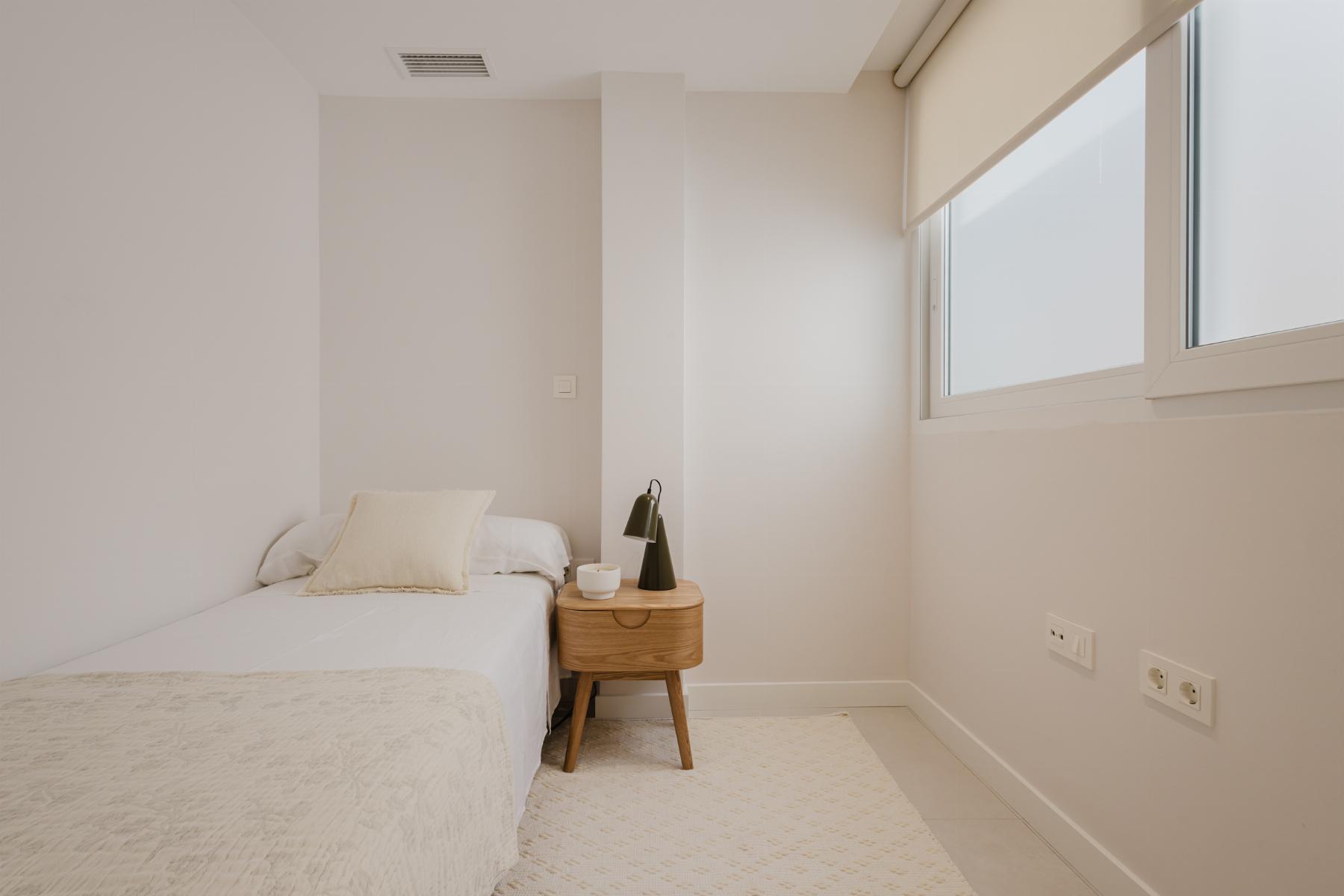 Minimal neutral bedroom with a single bed, a small wooden nightstand, black lamp, and a cup on the table beside a large window with blinds.