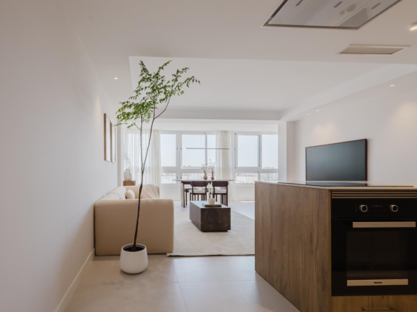 Bright minimalist living room with a tall potted plant, beige sofa, dining area by large windows, and a built-in oven.