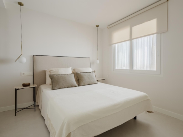 Minimalist bedroom with a beige upholstered bed, white bedding, two pendant lights, and a large window with a roller shade.