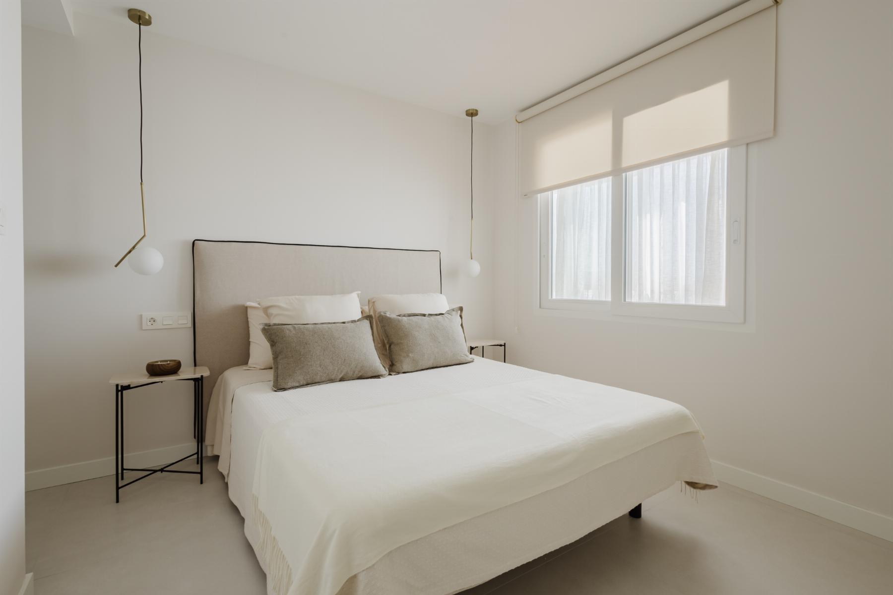 Minimalist bedroom with a beige upholstered bed, white bedding, two pendant lights, and a large window with a roller shade.