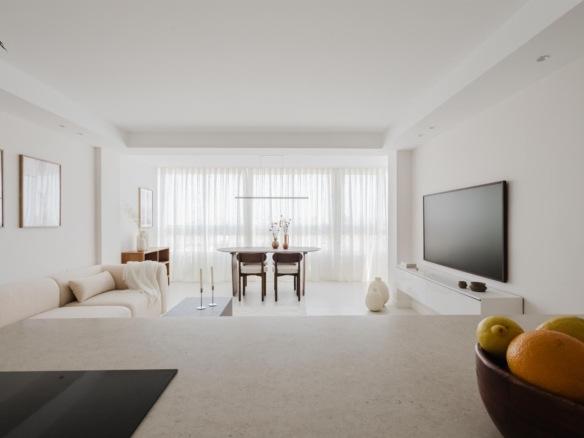 Bright minimalist living room with white furniture, dining area at the back, and a bowl of fruit on the counter in the foreground.