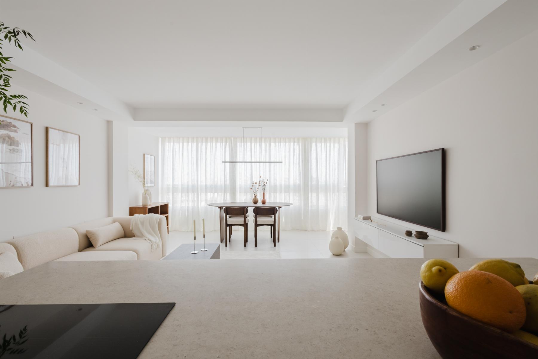 Bright minimalist living room with white furniture, dining area at the back, and a bowl of fruit on the counter in the foreground.