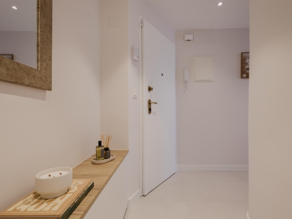 Modern hallway with white walls, a wooden console table, candle and reed diffuser, and a white front door in the center.