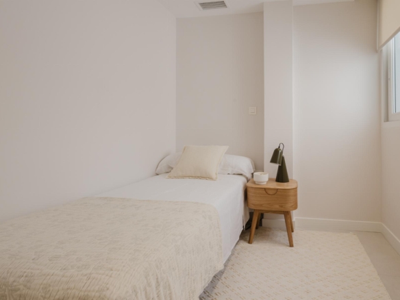 Minimalist bedroom with two twin beds pushed together, white bedding, and a beige pillow; a wooden nightstand with a green desk lamp and cup sits between beds; window on the right.