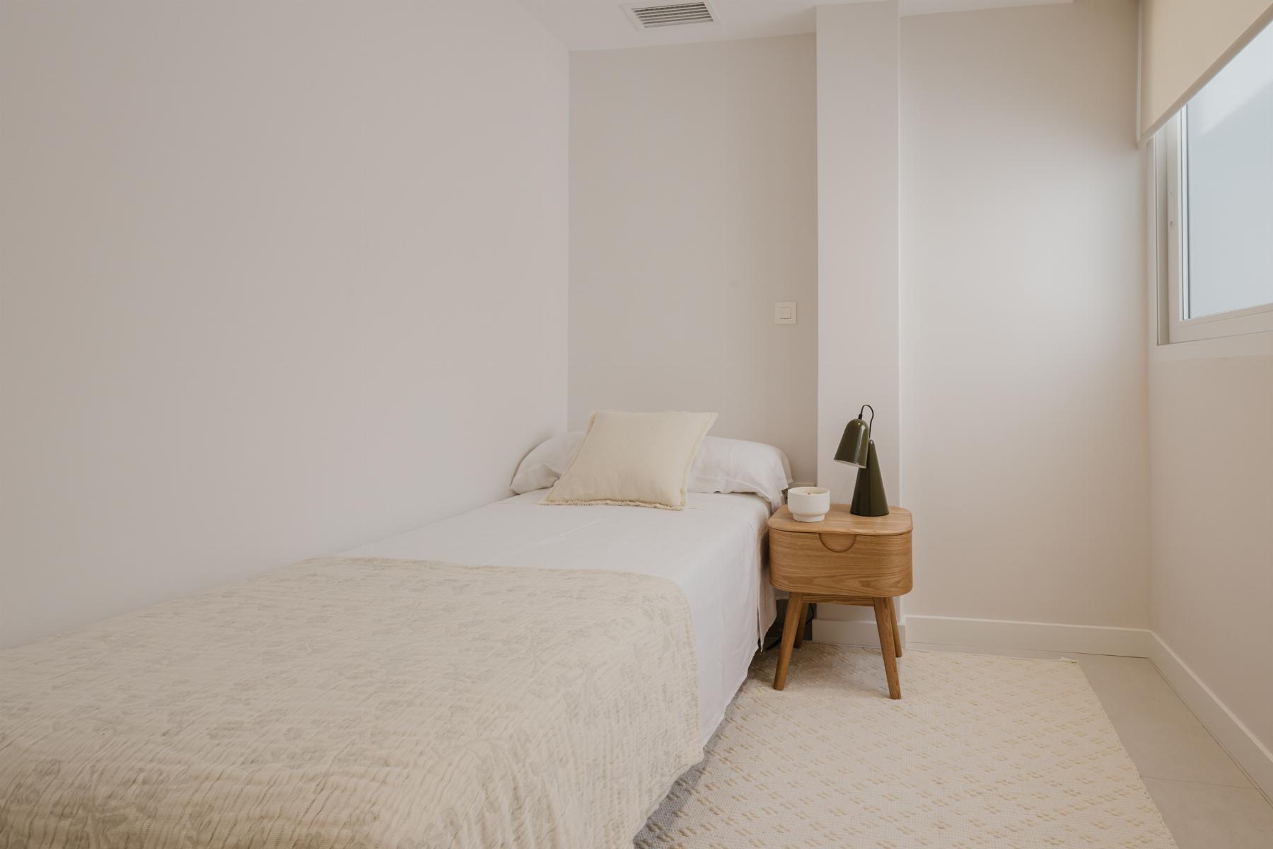 Minimalist bedroom with two twin beds pushed together, white bedding, and a beige pillow; a wooden nightstand with a green desk lamp and cup sits between beds; window on the right.