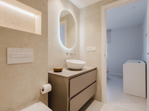 Modern bathroom with a backlit oval mirror above a vessel sink, beige tile walls, and a grey vanity with two drawers; toilet paper holder nearby. A doorway leads to a laundry area with a washing machine visible.