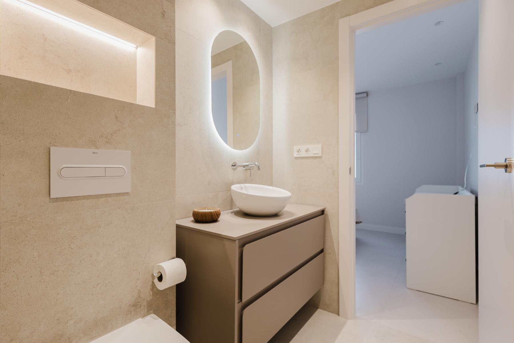 Modern bathroom with a backlit oval mirror above a vessel sink, beige tile walls, and a grey vanity with two drawers; toilet paper holder nearby. A doorway leads to a laundry area with a washing machine visible.