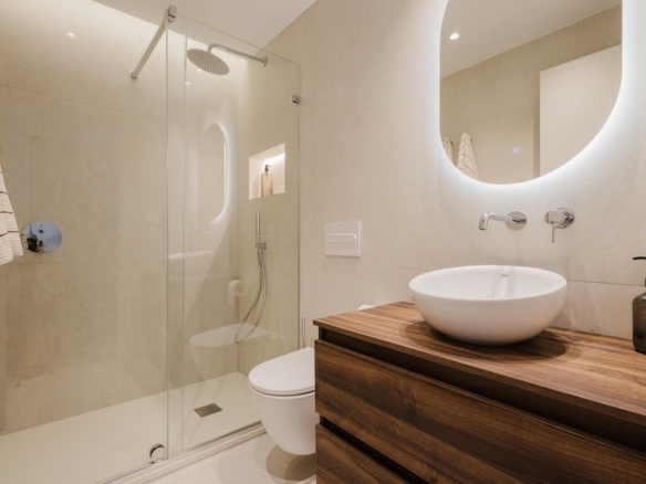 Modern beige tiled bathroom with glass shower enclosure, round white vessel sink on a wooden vanity, and a backlit curved mirror.